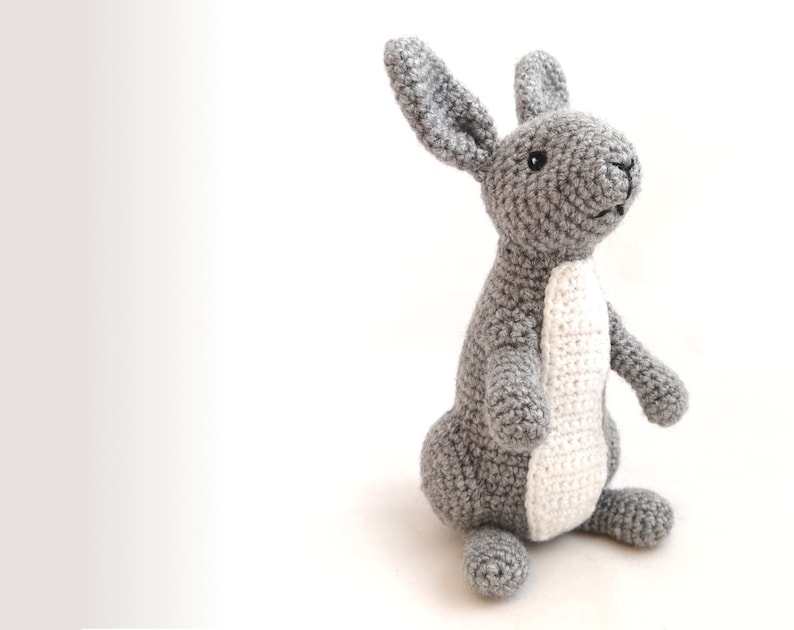 May include: A gray crocheted rabbit toy with white accents on its belly and ears. The rabbit is standing on a white surface.