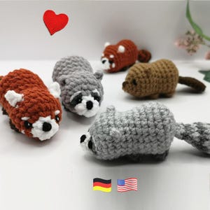Könnte beinhalten: Eine Sammlung von fünf kleinen, handgefertigten Häkeltierfiguren, darunter zwei graue Waschbären, ein roter Panda, ein brauner Biber und ein kleineres rostfarbenes Wesen. Ein rotes Herzsymbol und zwei kleine Flaggensymbole sind zu sehen.