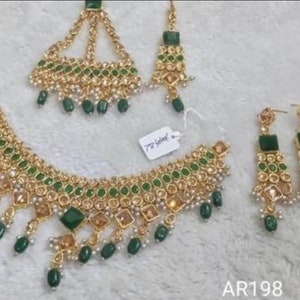 May include: A gold and green Indian bridal jewellery set featuring a necklace, earrings, and a headpiece. The necklace has a large, ornate pendant with green stones and pearls. The earrings are long and dangling, with green stones and pearls. The headpiece is a traditional Indian design with green stones and pearls.