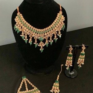 May include: A gold necklace, earrings, and a maang tikka set with green and clear stones. The jewellery is on a black background.
