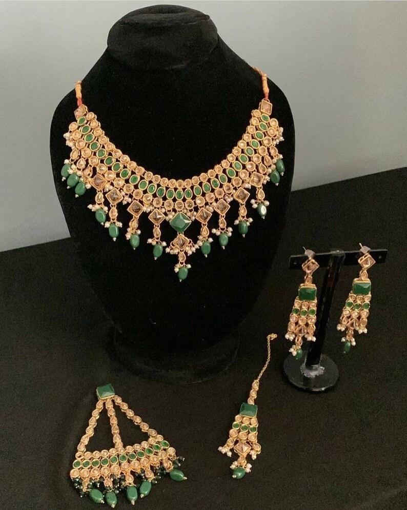May include: A gold necklace, earrings, and a maang tikka set with green and clear stones. The jewellery is on a black background.