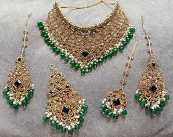 May include: A gold and green Indian bridal jewellery set with a necklace, earrings, and a tikka. The necklace features intricate floral designs and green and white beads. The earrings and tikka have matching designs.