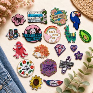 May include: A collection of embroidered patches featuring various designs including a stack of books, a toucan, a llama, a diamond, a sunflower, a safety pin with text, and circular patches with phrases like "Don't Give Up" and "I Am Enough."
