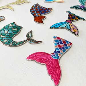 May include: Assortment of colourful embroidered iron-on patches featuring mermaid tails and a butterfly. The patches are made of fabric and have a variety of colours, including pink, blue, green, and orange.