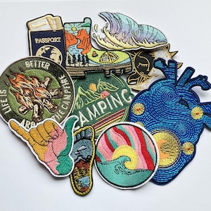 May include: A collection of embroidered iron-on patches featuring various designs, including a campfire, a wave, a heart, a camping scene, and a hand with a wave. The patches are colourful and feature a variety of textures.