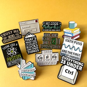Könnte beinhalten: Eine Sammlung von Emaille-Anstecknadeln mit verschiedenen Designs und Texten auf gelbem Hintergrund. Die Anstecknadeln zeigen Sätze wie "Trust Me I'm a Programmer", "Think Like a Proton" und "Math Puns". Weitere Anstecknadeln zeigen Bücher, eine Tabelle und eine Computertaste.