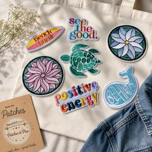 Può includere: Una collezione di toppe ricamate termoadesive che include una tavola da surf con il testo beach bum, il testo see the good, una tartaruga marina verde, un fiore rosa, un fiore blu, il testo positive energy e una balena blu con il testo BE HAPPY.