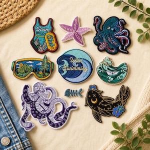 Op de afbeelding: Een verzameling van negen geborduurde patches met diverse ontwerpen, waaronder een badpak met de tekst Swim Wild Club, een paarse zeester, een blauwe octopus, een skibril met een berglandschap, een ronde patch met de tekst Wild Swimming, een papieren bootje op golven, een paarse octopus, een visgraat en een zwarte vis met gouden hemelse details.