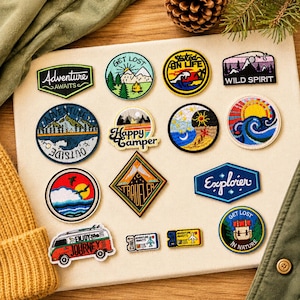 May include: A collection of embroidered patches featuring outdoor and travel themes. Designs include mountains, forests, waves, a camper van, and text such as Adventure Awaits, Get Lost, Wild Spirit, Happy Camper, Traveler, Explorer, and Enjoy the Journey.