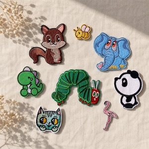 May include: A collection of embroidered fabric patches featuring a brown squirrel, a yellow and pink bee, a blue elephant, a green dinosaur, a green caterpillar, a black and white panda, a teal cat face, and a pink flamingo on a beige fabric.
