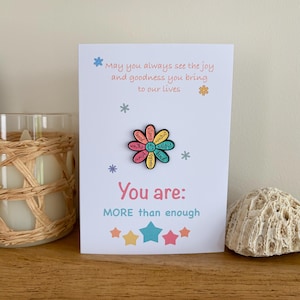 May include: A white greeting card featuring a colorful flower-shaped enamel pin at the center. The card text reads: May you always see the joy and goodness you bring to our lives. You are: MORE than enough. The pin petals contain positive words.