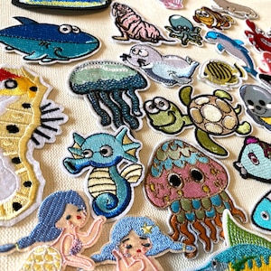Könnte beinhalten: Eine Sammlung bunter, gestickter Bügelbilder mit verschiedenen Meeresbewohnern, darunter ein blauer Hai, ein gelbes Seepferdchen, eine grüne Schildkröte, eine rosa Qualle und ein blaues Seepferdchen.
