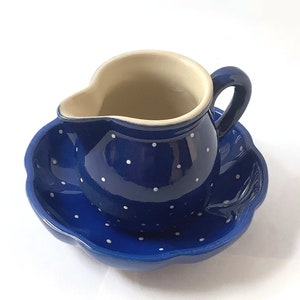 May include: Blue and white ceramic creamer with a matching saucer. The creamer has a white interior and a blue exterior with white polka dots. The saucer has a scalloped edge.