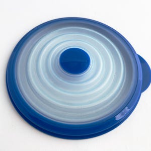 Könnte beinhalten: Ein blau-weißer Kunststoffdeckel mit kreisförmigem Design. Der Deckel hat einen dunkelblauen Rand und eine hellere blaue Mitte mit konzentrischen Ringen. Ein kleiner, dunkelblauer Griff befindet sich an der Seite. Die Mitte hat den Markennamen.