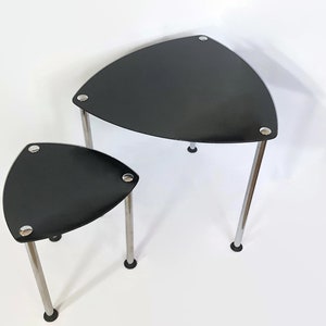 Op de afbeelding: Een set van twee zwarte driehoekige nesttafels met chromen poten. De tafels zijn van glas en hebben een modern ontwerp.