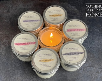 May include: A collection of six soy wax candles in glass jars with silver lids. The candles are arranged in a circle on a dark gray surface. Each candle has a label with the name of the scent and the text "Nothing Less Than Home, LLC". The scents are Blackberry Sage, Honeysuckle, Raspberry Vanilla, Raspberry Vanilla, Currant & Absinthe, and Honeysuckle. The text "NOTHING Less Than HOME" is written in white letters on a black background above the candles.