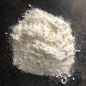 May include: A close-up of a pile of white laundry detergent powder with small white flakes and white plastic beads.