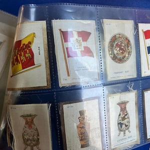 Könnte beinhalten: Eine Sammlung von Vintage-Sammelkarten in einer Plastikhülle. Die Karten zeigen Illustrationen von Flaggen, Keramikkunst und Vasen. Zu den Flaggen gehören Wales, Dänisch-Westindien und Holland. Die Karten sind in verschiedenen Farben gehalten.