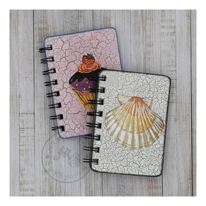 Pode incluir: Dois pequenos cadernos de espiral com capas decorativas. Um caderno tem uma capa rosa com um efeito de pintura rachada e um cupcake com cobertura roxa e um morango no topo. O outro caderno tem uma capa branca com um efeito de pintura rachada e uma concha com listras amarelas e laranja.