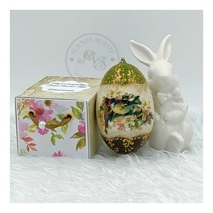 Könnte beinhalten: Ein dekoratives Osterei mit einem floralen und Vogelmotiv, bemalt in Grün-, Braun- und Goldtönen. Das Ei wird in einer Schachtel mit Blumenmuster und einem weißen Porzellanhasen präsentiert.