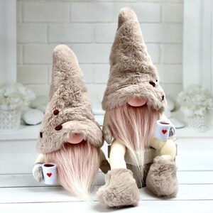 May include: Two plush gnomes with tall, furry hats and long pink beards. Each gnome holds a small white mug with a red heart. The hats are adorned with brown coffee bean accents. The gnomes are a light brown colour.