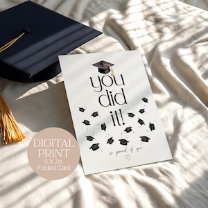 Może przedstawiać: Karta gratulacyjna z napisem "You did it!" i spadającymi biretami. Karta jest drukiem cyfrowym, złożonym 12,7 x 17,8 cm. Czarne nakrycie głowy i chwost w tle.