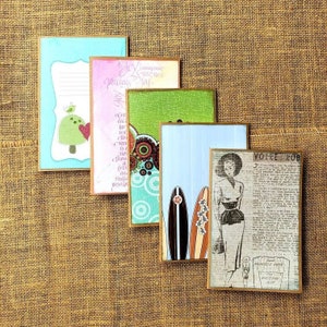 Peut inclure: Cinq cartes différentes avec des motifs variés. Les cartes sont toutes rectangulaires et ont une bordure brune. Les motifs comprennent une carte bleue et verte avec un oiseau et un champignon, une carte rose et blanche avec un motif floral, une carte verte et brune avec un motif circulaire, une carte bleue avec deux planches de surf et une carte brune avec une femme en robe et le texte "VOTRE ROB".