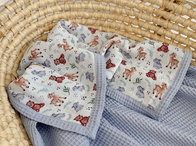 May include: A baby blanket with a white background and a pattern of brown deer, red teddy bears, and gray hippos. The blanket has a blue waffle weave texture and a blue border. The blanket is in a wicker basket.