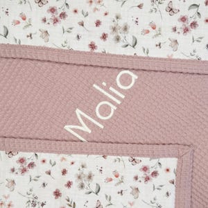 May include: A baby blanket with a floral pattern and a textured, pink waffle weave. The blanket features the name "Malia" in white script. The floral pattern includes butterflies and flowers. The blanket is a soft, cozy item.
