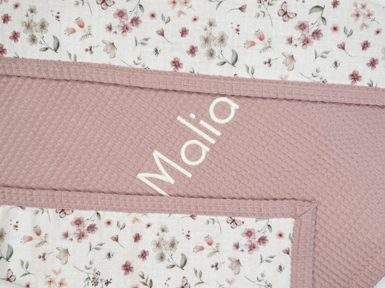 May include: A baby blanket with a floral pattern and a textured, pink waffle weave. The blanket features the name "Malia" in white script. The floral pattern includes butterflies and flowers. The blanket is a soft, cozy item.