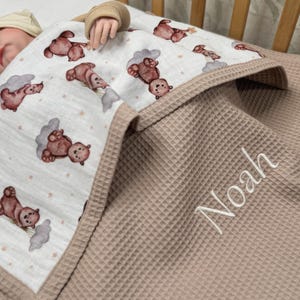 May include: A baby blanket with a teddy bear pattern and the name "Noah" embroidered in white. The blanket has a waffle texture in a neutral beige color, with a patterned top layer featuring brown teddy bears and clouds.