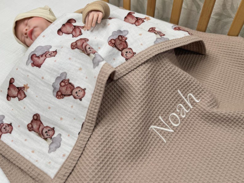 May include: A baby blanket with a teddy bear pattern and the name "Noah" embroidered in white. The blanket has a waffle texture in a neutral beige color, with a patterned top layer featuring brown teddy bears and clouds.