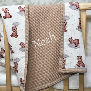 May include: A baby blanket with a waffle-textured center in a light brown color. The edges feature a white fabric with a repeating pattern of brown teddy bears on clouds. The name "Noah" is embroidered on the blanket.