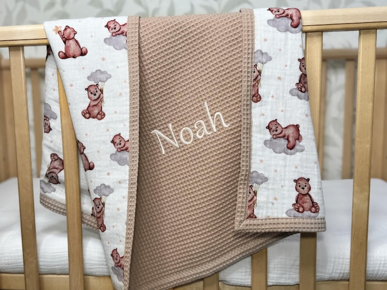 May include: A baby blanket with a waffle-textured center in a light brown color. The edges feature a white fabric with a repeating pattern of brown teddy bears on clouds. The name "Noah" is embroidered on the blanket.