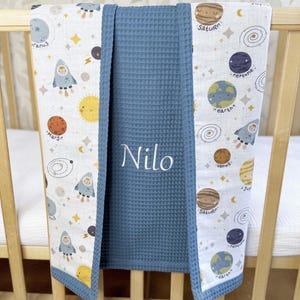 May include: A baby blanket with a space theme, featuring planets, rockets, and stars on a white background. The blanket has a blue waffle-weave center with the name "Nilo" embroidered in white. The edges are trimmed in blue.
