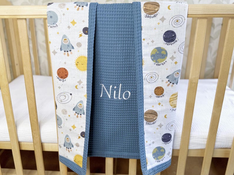 May include: A baby blanket with a space theme, featuring planets, rockets, and stars on a white background. The blanket has a blue waffle-weave center with the name "Nilo" embroidered in white. The edges are trimmed in blue.