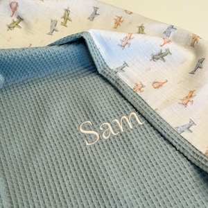 May include: A baby blanket with a blue waffle weave texture and a white airplane print. The name "Sam" is embroidered in white thread on the blue fabric. The blanket has a soft, cozy appearance.