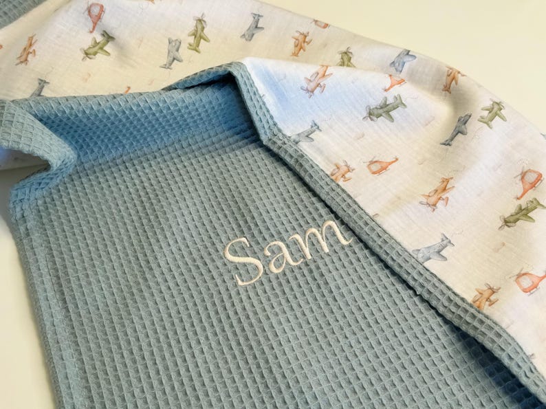 May include: A baby blanket with a blue waffle weave texture and a white airplane print. The name "Sam" is embroidered in white thread on the blue fabric. The blanket has a soft, cozy appearance.