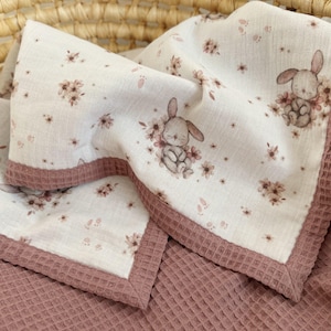 May include: A baby blanket with a white floral and bunny print, framed by a mauve waffle-weave border. The blanket is folded, revealing the soft, textured fabric and the delicate illustrations of bunnies and flowers.