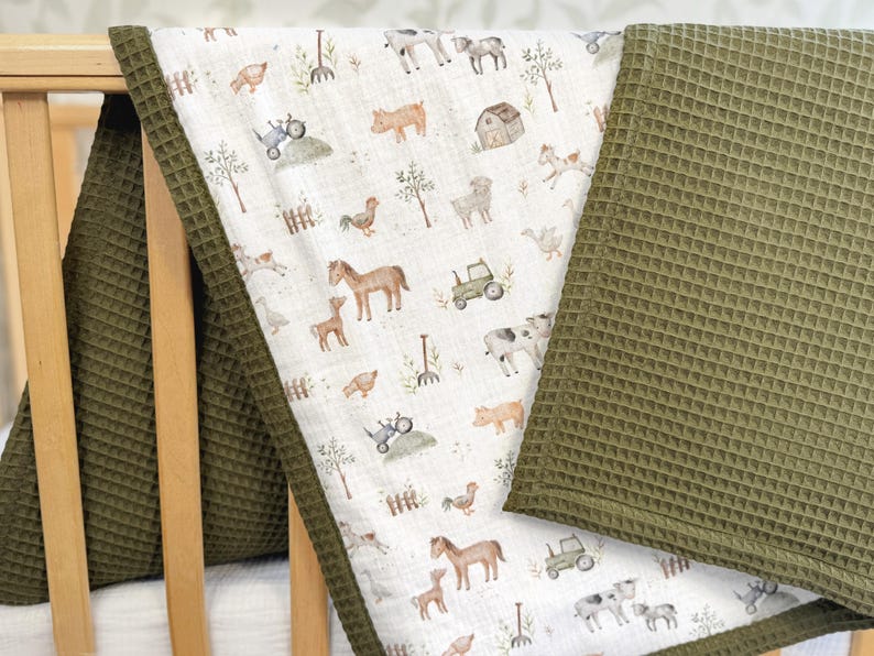May include: A baby blanket with a farm animal pattern and a green waffle weave border. The blanket features illustrations of horses, cows, pigs, and tractors on a white background. The border and a separate blanket are olive green.