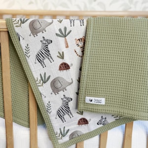 May include: A baby blanket with a safari animal print and a solid green waffle weave. The patterned side features elephants, zebras, and turtles. The blanket is draped over a wooden crib.