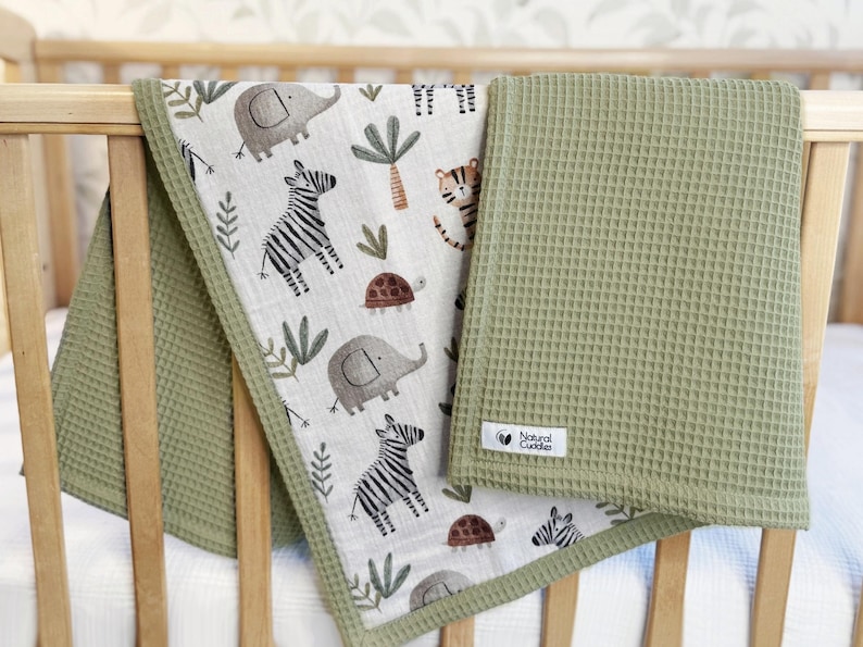 May include: A baby blanket with a safari animal print and a solid green waffle weave. The patterned side features elephants, zebras, and turtles. The blanket is draped over a wooden crib.