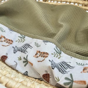 May include: A baby blanket with a white border featuring a jungle animal print of tigers, zebras, and turtles. The blanket has a green waffle weave texture and is folded inside a wicker basket.