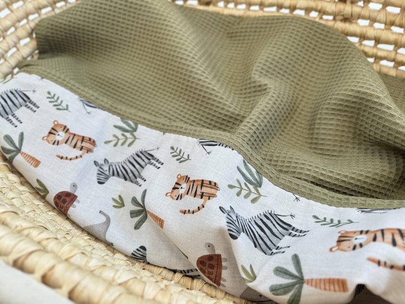 May include: A baby blanket with a white border featuring a jungle animal print of tigers, zebras, and turtles. The blanket has a green waffle weave texture and is folded inside a wicker basket.