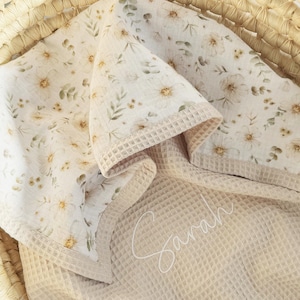 May include: A beige baby blanket with a floral pattern and a waffle-weave border. The blanket is folded in a woven basket. The floral pattern includes white flowers and green leaves. The name "Sarah" is embroidered on the blanket.