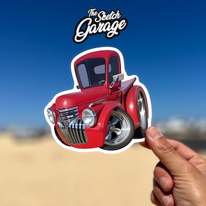 May include: Red cartoon pickup truck sticker with "The Sketch Garage" text above it. The truck has a white outline and is being held by a hand.