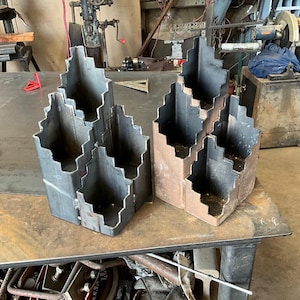 Puede incluir: Cuatro estructuras metálicas con diseño escalonado, probablemente para construcción o uso industrial. Dos son de color gris plateado, dos tienen un tono marrón. Están sobre una superficie metálica en un taller.