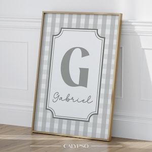 Può includere: Opera d'arte da parete incorniciata con sfondo a motivo gingham grigio. L'opera presenta un centro bianco con la lettera "G" grigia e il nome "Gabriel" in corsivo. La cornice è in legno chiaro.