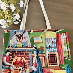 Op de afbeelding: Een rechthoekige tote bag met twee beige stoffen hengsels, voorzien van een kleurrijke naaldwerksc&egrave;ne van een persoon zittend bij een bakstenen open haard, een ander persoon die ernaast staat en een hond in een gedetailleerd interieur.