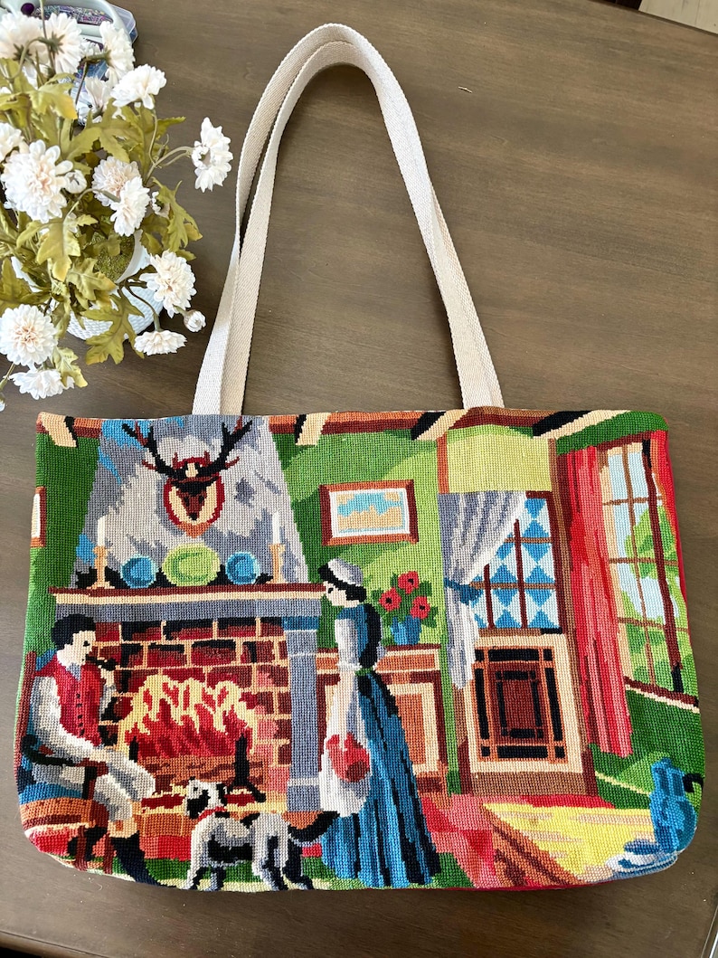 Op de afbeelding: Een rechthoekige tote bag met twee beige stoffen hengsels, voorzien van een kleurrijke naaldwerksc&egrave;ne van een persoon zittend bij een bakstenen open haard, een ander persoon die ernaast staat en een hond in een gedetailleerd interieur.